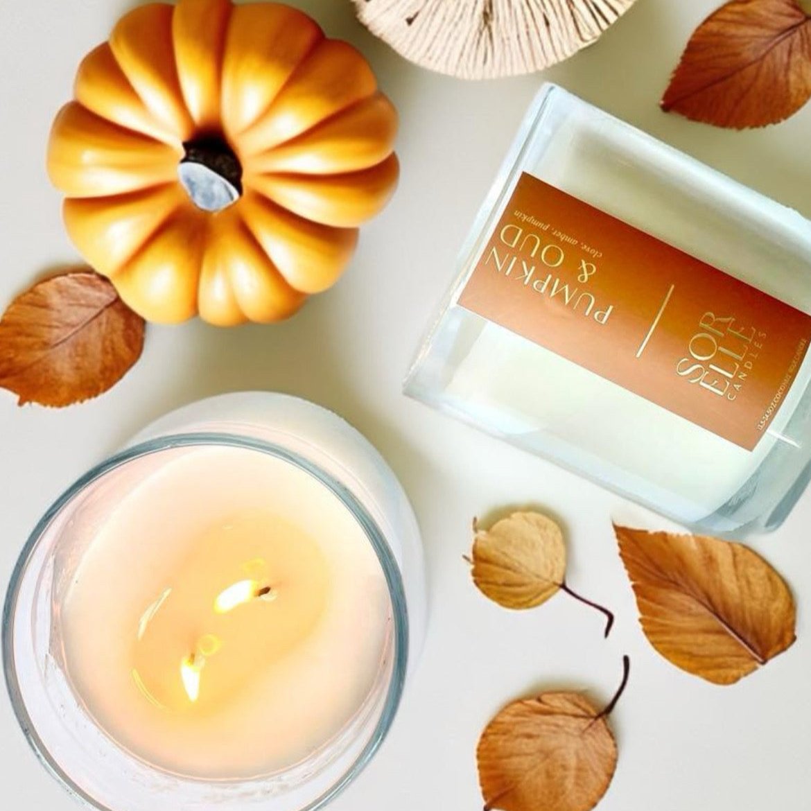 Flat lay of Pumpkin Oud candle with dried spices, mini pumpkins, and fall foliage on linen fabric