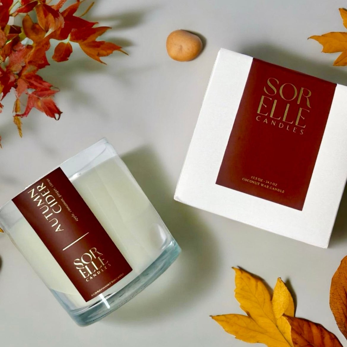 A coconut wax candle in a clear glass jar with a red label, placed next to its white box with red branding, surrounded by autumn leaves and a cinnamon stick.